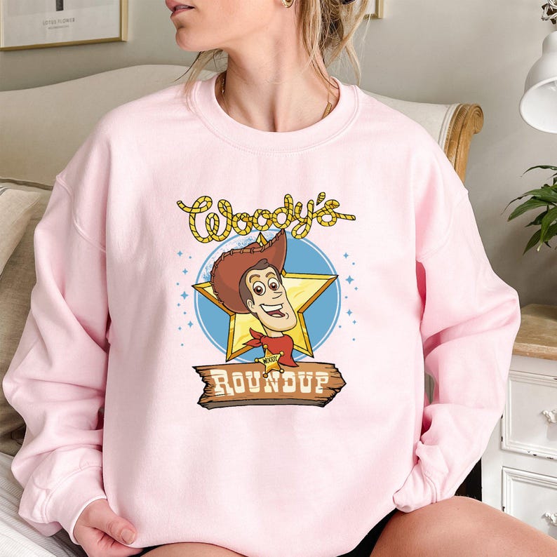 Peut inclure: Sweat-shirt rose avec une image de dessin anim&eacute; de Woody de Toy Story. Le graphique repr&eacute;sente le visage de Woody et le texte "Woody's Roundup".