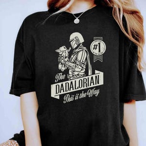 K&ouml;nnte beinhalten: Schwarzes T-Shirt mit einer wei&szlig;en Grafik eines Mandalorianer-Charakters, der ein Baby Yoda h&auml;lt. Der Text "The Dadalorian This is the Way" ist unter dem Bild gedruckt. Ein wei&szlig;er Kreis mit dem Text "Certified #1 in the Galaxy" befindet sich in der oberen rechten Ecke.