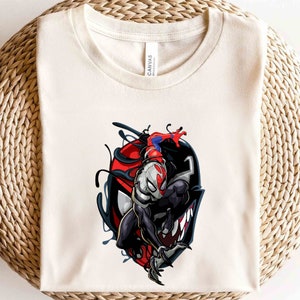 Puede incluir: Una camiseta blanca con un gr&aacute;fico de Venom y Spider-Man. Venom est&aacute; en primer plano, con Spider-Man detr&aacute;s de &eacute;l. Venom es negro con ojos y dientes blancos, y Spider-Man es rojo y azul.