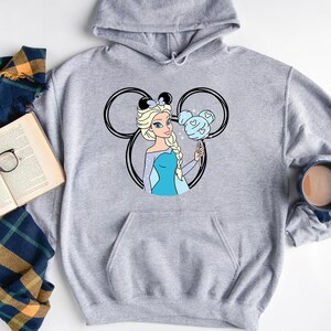 Puede incluir: Sudadera gris con capucha con una ilustraci&oacute;n en blanco y negro de una princesa Disney con cabello azul sosteniendo un dulce con forma de Mickey Mouse azul.