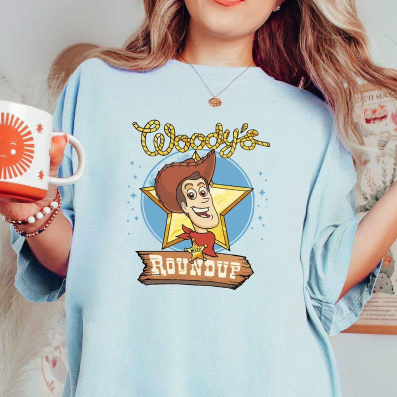 Peut inclure: T-shirt bleu clair avec une image de dessin anim&eacute; de Woody de Toy Story. L'image montre Woody avec son chapeau de cowboy, son gilet et son badge de sh&eacute;rif embl&eacute;matiques. Le texte "Woody's Roundup" est imprim&eacute; sous l'image.