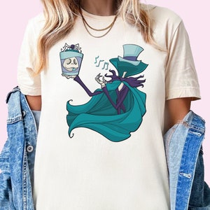 Puede incluir: Una camiseta blanca con una ilustraci&oacute;n de dibujos animados de color turquesa y morado de un esqueleto con un sombrero de copa y una capa, sosteniendo un frasco con una calavera en su interior. El esqueleto est&aacute; cantando y notas musicales flotan a su alrededor.