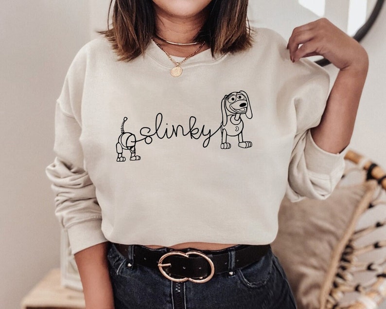 May include: A cream-colored sweatshirt with a black outline of Slinky Dog from the Toy Story franchise. The text "Slinky" is written in cursive above the dog.