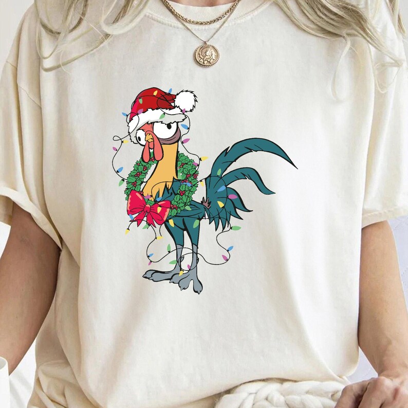 May include: A white t-shirt with a cartoon rooster wearing a Santa hat and holding a Christmas wreath with lights around it.