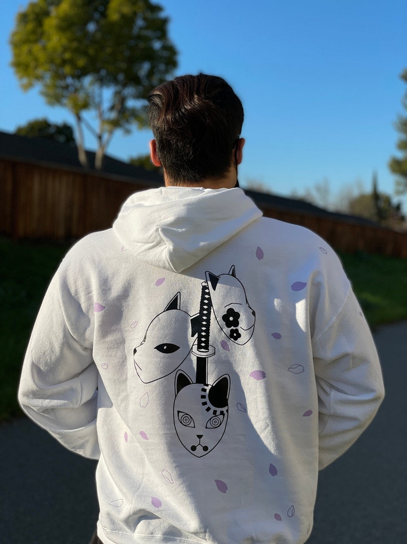 May include: White hoodie featuring a graphic design of three anime masks and a sword. The design is in black and white, with purple accents of falling petals. The hoodie is a comfortable, casual style.