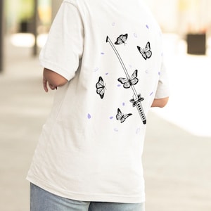 Pode incluir: Camiseta branca com um gr&aacute;fico preto e branco de uma espada e borboletas. As borboletas voam ao redor da espada, que est&aacute; no centro do design.