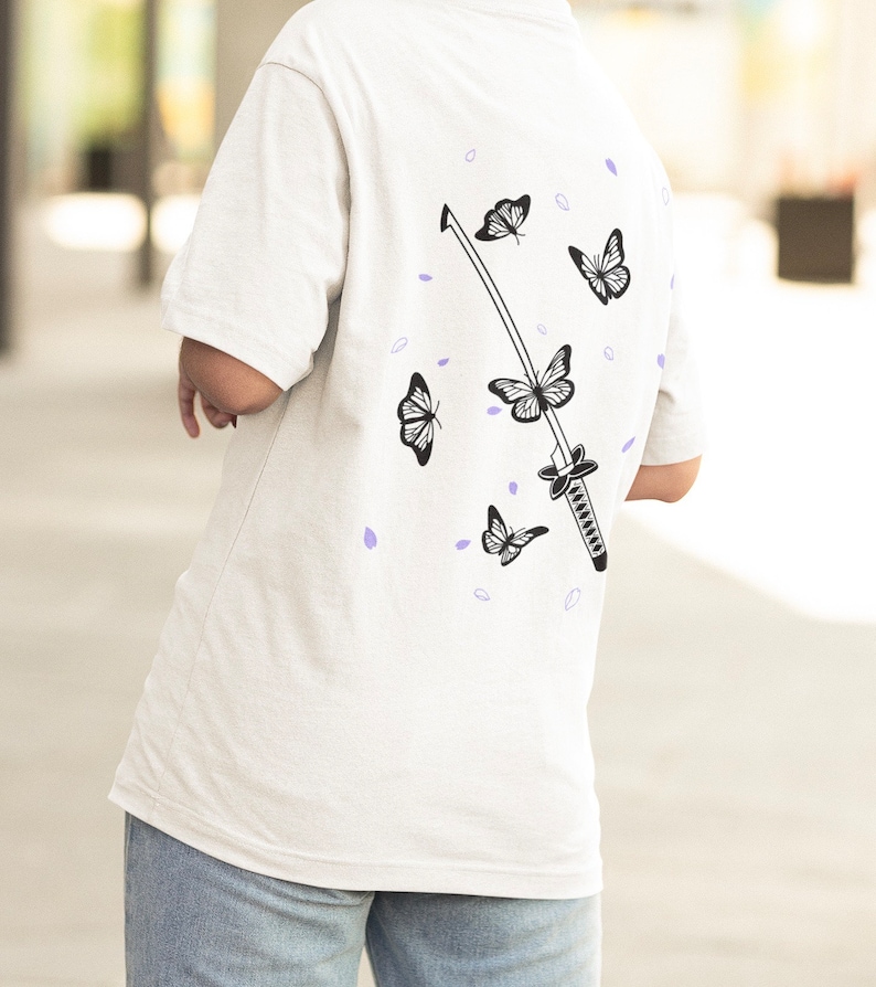Pode incluir: Camiseta branca com um gr&aacute;fico preto e branco de uma espada e borboletas. As borboletas voam ao redor da espada, que est&aacute; no centro do design.
