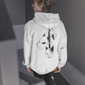 May include: White hoodie with a black graphic of three stylized masks and a sword on the back. The masks have different designs, and the hoodie has a hood and long sleeves. The person is wearing black pants.