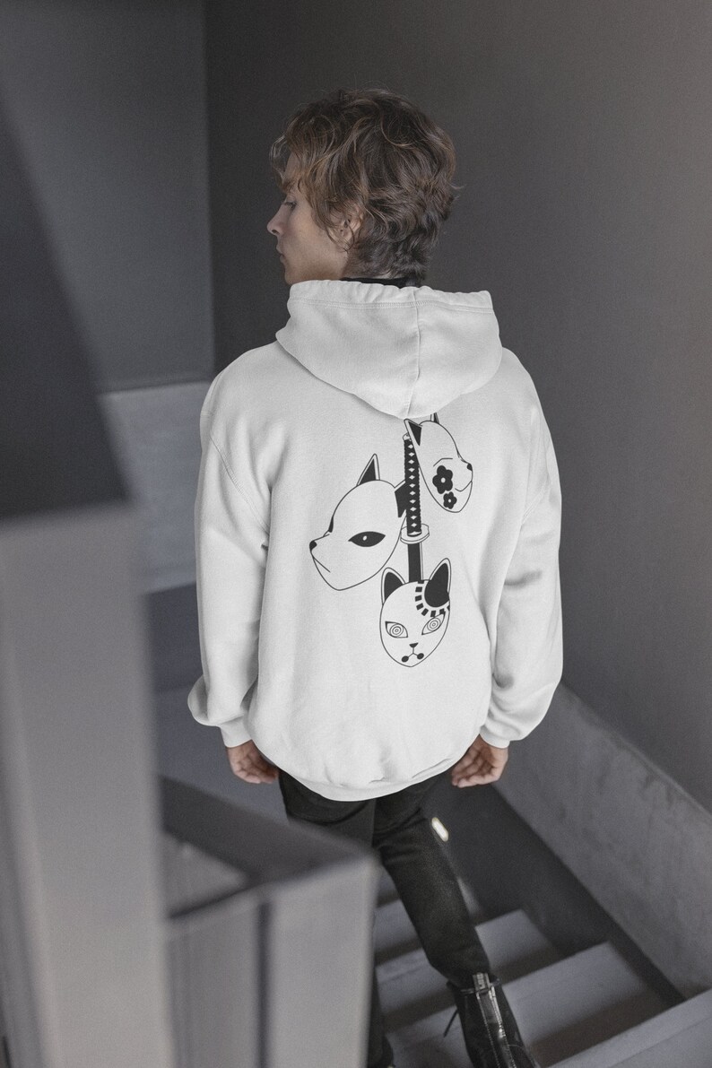 May include: White hoodie with a black graphic of three stylized masks and a sword on the back. The masks have different designs, and the hoodie has a hood and long sleeves. The person is wearing black pants.