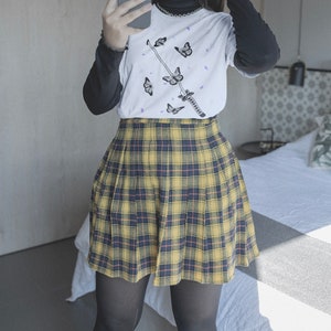 Pode incluir: Uma saia plissada xadrez amarela e preta com uma camiseta branca com uma espada preta e borboletas roxas.