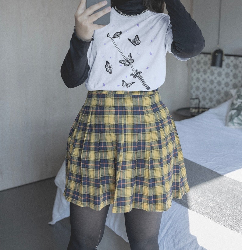 Pode incluir: Uma saia plissada xadrez amarela e preta com uma camiseta branca com uma espada preta e borboletas roxas.