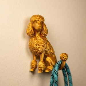 May include: A golden-colored poodle-shaped wall hook. The hook is designed to hold items and features a detailed sculpture of a poodle. A blue and black rope is hanging from the hook. The hook is mounted on a beige wall.