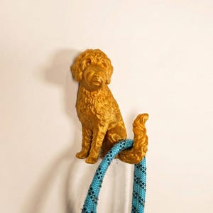 May include: A golden-colored dog-shaped wall hook. The hook is designed to hold a leash, which is blue with a black pattern. The dog is sitting and has a curly coat. The hook is mounted on a white wall.