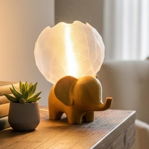 May include: A decorative table lamp featuring a yellow elephant base and a white, spherical shade. The lamp sits on a wooden table next to a small potted succulent and a stack of books. The lamp's shade emits a warm, inviting glow.