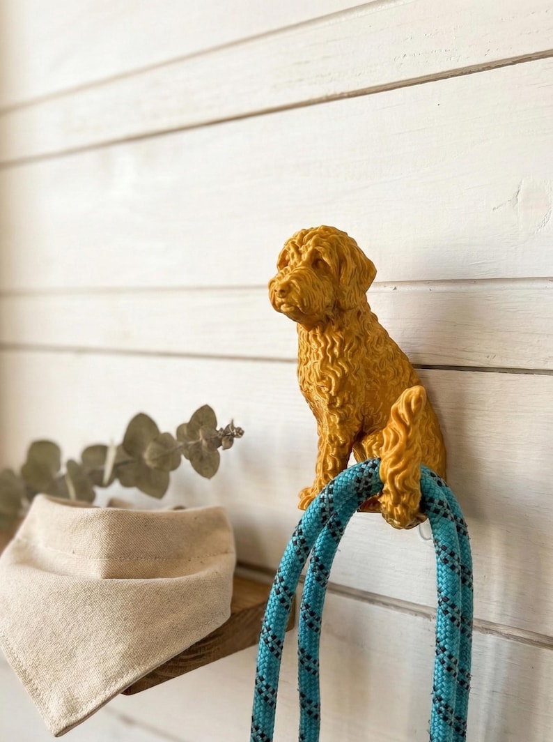 May include: A golden-colored dog-shaped wall hook holding a blue and black rope. The hook is mounted on a white wooden wall. A tan dog bandana and eucalyptus sprig are visible on a wooden shelf below.