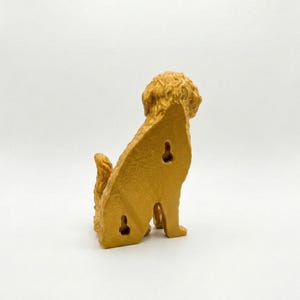 May include: A golden-colored dog-shaped wall hook, viewed from the back, with a textured, curly coat and tail. The dog is sitting, with three keyhole-shaped openings on its back for mounting. The background is plain white.