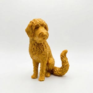 May include: A golden-colored figurine of a dog in a sitting position with a curly tail. The dog has a textured coat and is set against a white background. The figurine is a decorative item.