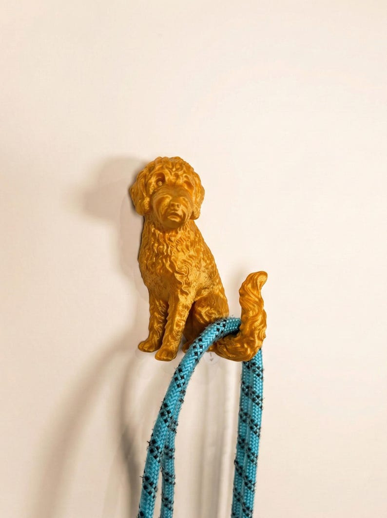 May include: A golden-colored dog-shaped wall hook. The hook is designed to hold a leash, which is blue with a black pattern. The dog is sitting and has a curly coat. The hook is mounted on a white wall.