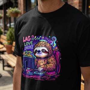 May include: Black t-shirt featuring a colorful graphic of a sloth working on a computer. The text "LAG IS MY LOVE LANGUAGE" is above the sloth. The sloth is sitting in a pink chair with a purple backpack.