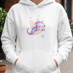 Könnte beinhalten: Weißer Kapuzenpullover mit einer Illustration eines Cartoon-Axolotls in Pastellfarben. Das Axolotl hat Flügel und den Text "Float & Relax". Der Hoodie hat eine Kapuze mit Kordelzug und eine Vordertasche. Das Design ist skurril und verspielt.