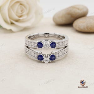May include: A silver-toned ring guard set featuring two bands with milgrain edges. Each band is adorned with a central clear round-cut stone flanked by two deep blue round-cut stones, with additional small clear stones along the shanks.