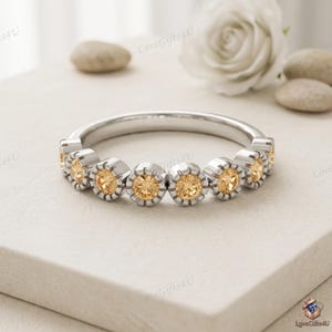 Op de afbeelding: Een zilverkleurige ring met een reeks van zeven rondgeslepen, lichtoranje edelstenen. Elke steen is individueel gezet in een ronde, gekartelde metalen zetting, wat zorgt voor een doorlopend en uniform ontwerp aan de voorzijde van de ring.