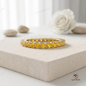 Könnte beinhalten: Ein kreisförmiger Eternity-Ring mit einer durchgehenden Reihe von rund geschliffenen, leuchtend gelben Edelsteinen, die in ein poliertes Metallband gefasst sind. Der Ring liegt horizontal und zeigt die einheitliche Größe und Farbe.