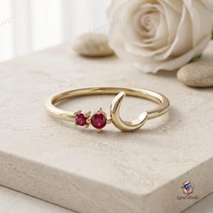 Puede incluir: Un anillo de tono dorado que presenta una forma de media luna y dos pequeñas piedras preciosas rojas redondas colocadas una al lado de la otra. La banda es delgada y pulida, con la luna y las piedras situadas en la curva superior del anillo.