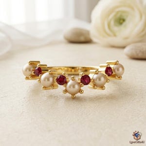 May include: A gold ring featuring a V-shaped band set with alternating round white pearls and small round red rubies. The pearls and rubies are held in place by gold prongs, creating a symmetrical arrangement across the top of the band.