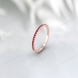 Puede incluir: Anillo de oro rosa con una banda de pequeñas gemas rojas vibrantes. El anillo se presenta sobre un fondo blanco suave, resaltando el diseño delicado y el brillo de las gemas. Una pieza de joyería sencilla y elegante.