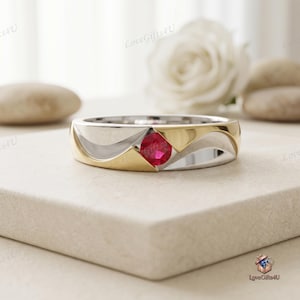 Può includere: Un anello in metallo bicolore caratterizzato da una fascia color argento lucido con accenti color oro e una singola pietra preziosa rotonda di colore rosso vivace incastonata centralmente in un'apertura triangolare sulla parte anteriore.