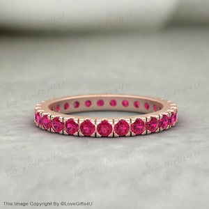 Pode incluir: Um anel de eternidade em ouro rosa com uma fileira contínua de pedras preciosas rosa vibrantes. As pedras preciosas são de corte redondo e são colocadas próximas umas das outras, criando uma faixa completa de cor. O anel é exibido em um fundo neutro.