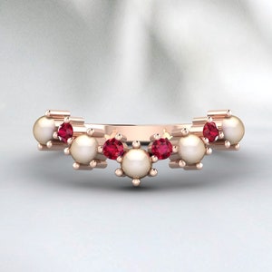 Puede incluir: Un anillo de oro rosa con perlas y gemas rojas alternadas. El anillo tiene un diseño curvo, con las gemas y perlas dispuestas en un patrón decorativo. El anillo es una joya.