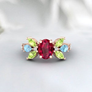 May include: Rose gold ring featuring an oval-cut ruby center stone. The ring is adorned with pear-shaped peridot and aquamarine stones, along with small diamonds. The design is symmetrical, with the gemstones arranged around the central ruby.