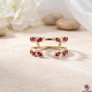 Puede incluir: Un anillo dorado con múltiples piedras preciosas de color rubí, talladas en forma de marquesa. El anillo tiene un diseño único, con dos bandas curvas que parecen abrazar otro anillo. Las piedras preciosas están espaciadas uniformemente a lo largo de las bandas.