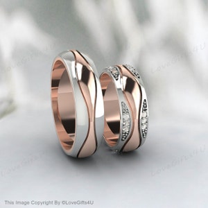 Puede incluir: Dos anillos de boda de dos tonos. Uno presenta un diseño de onda con diamantes incrustados. Los anillos tienen una combinación de colores plateado y oro rosa. Se muestran sobre un fondo blanco.