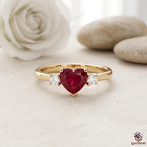 Op de afbeelding: Een gouden ring met een centrale hartvormige rode edelsteen, geflankeerd door twee kleine ronde heldere stenen. De band is van gepolijst geelgoud. Het ontwerp is symmetrisch met de hartvormige steen in het midden en een ronde steen aan weerszijden.