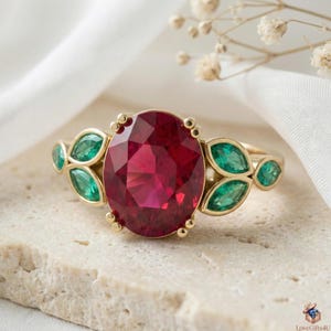 Peut inclure: Une bague en or ornée d'un grand rubis ovale facetté en son centre. La bague est ornée d'émeraudes taille émeraude de chaque côté, disposées en forme de feuilles. La bague est présentée sur une surface en pierre claire.