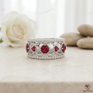 Peut inclure: Une bague en métal argenté ornée d'une rangée centrale de pierres rouges rondes serties dans une structure en treillis complexe. Les bords supérieur et inférieur de l'anneau sont bordés de petites pierres claires pavées, créant un design détaillé.