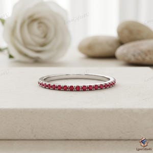 May include: A thin, silver-toned metal band ring featuring a continuous row of small, round, vibrant red gemstones set along the outer circumference. The ring has a polished finish and a slim profile, showcasing a uniform design of red stones.