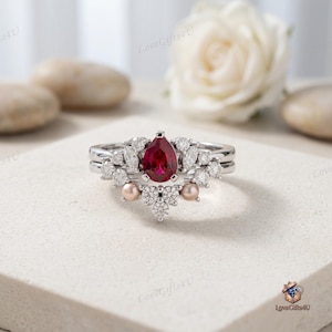 Puede incluir: Un anillo plateado con una piedra roja central en forma de pera, rodeada de pequeñas piedras claras redondas y de talla marquesa, y dos pequeñas perlas redondas de color rosa pálido integradas en el diseño decorativo del anillo.