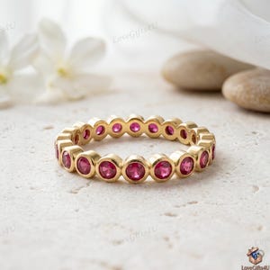 May include: A gold eternity band featuring a continuous row of round, bezel-set vibrant pink gemstones. The ring has a polished metallic finish and a uniform circular design, showcasing a series of identical stones set securely in gold metal.