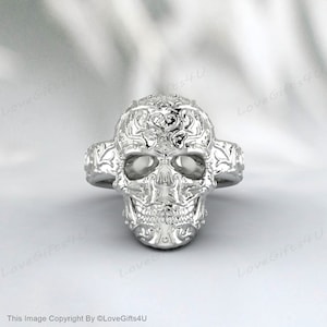 Puede incluir: Un anillo de calavera plateado con intrincados detalles florales y de volutas. El anillo presenta un diseño de calavera detallado con cuencas de los ojos abiertas y una frente detallada. La banda del anillo tiene un diseño decorativo a cada lado.