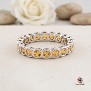 Könnte beinhalten: Ein silberfarbener Eternity-Ring mit einer durchgehenden Reihe von rund geschliffenen, goldgelben Edelsteinen, die in einzelnen kreisförmigen Metallfassungen sitzen. Der Ring zeigt ein einheitliches Design mit polierten Metallrändern.