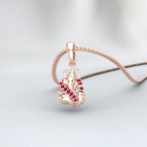 May include: A rose gold pendant necklace featuring a detailed Ganesh design. The pendant is adorned with small, round, red gemstones. The necklace has a delicate chain and is displayed on a light gray background.