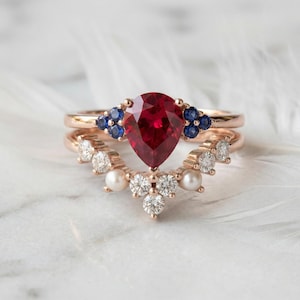 May include: Two rose gold rings. The top ring features a large, pear-shaped, deep red gemstone, accented with small blue stones. The bottom ring has a curved design with diamonds and pearls. The rings are set against a white background.