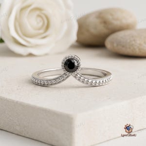 May include: A silver chevron-shaped ring featuring a single round-cut black gemstone set in the centre. The band is decorated with a delicate milgrain beaded texture along the entire outer edge, creating a detailed and structured appearance.