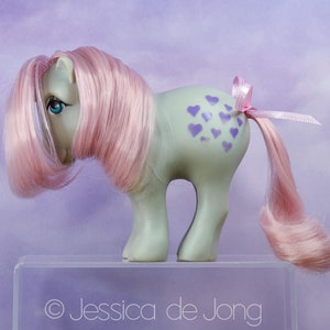May include: A vintage toy pony with a light green body, pink hair, and blue eyes. The pony has purple heart-shaped markings on its flank and a pink ribbon tied to its tail. The pony is displayed on a clear stand against a purple background.