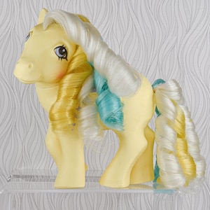 Könnte beinhalten: Ein gelbes Vintage-Spielzeugpferd mit weißen, gelben und blauen Locken. Das Pony hat blaue Augen und rosige Wangen und steht auf einem transparenten Acrylregal. Ein Sammlerspielzeug.