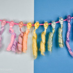 May include: A collection of colorful, curly doll hair extensions in shades of pink, purple, yellow, and green, attached to a pink string. The background is split between light gray and blue.